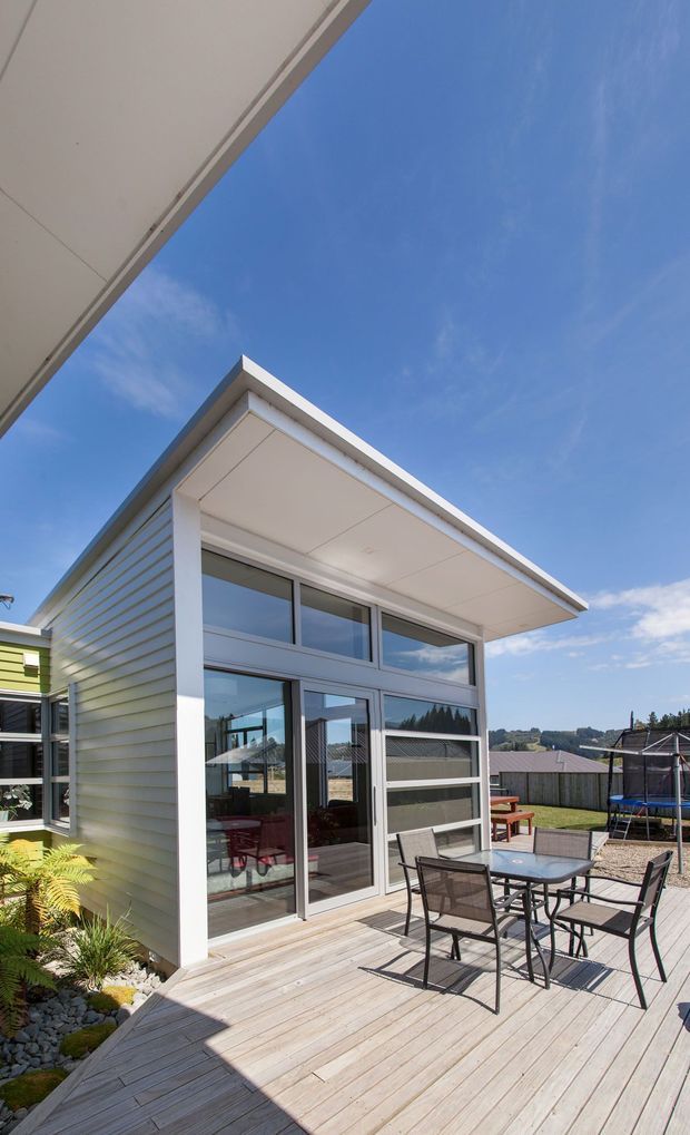Bright exterior view of a contemporary single-storey home extension or pavilion opening onto a timber deck. The structure has white horizontal weatherboard cladding, extensive floor-to-ceiling glazing with aluminium frames, a broad flat projecting roof, and a furnished outdoor dining area. A fenced suburban landscape and clear blue sky are visible beyond.