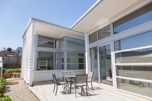 Bright exterior view of a contemporary residential extension or courtyard zone, featuring white horizontal weatherboard cladding, extensive aluminium-framed glazing, flat roof forms, and a timber-look deck with outdoor dining furniture. The image is taken from outside and primarily shows the building facade and covered patio area.