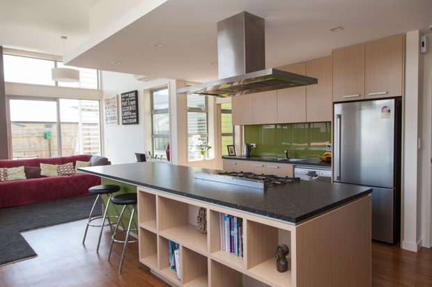 Bright open-plan kitchen and living area with a large dark stone island featuring integrated shelving and bar seating. Light timber cabinetry, a glossy green backsplash, stainless-steel rangehood and appliances, timber flooring, and extensive glazing create a contemporary family-oriented interior.