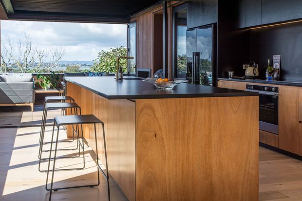 A contemporary open-plan kitchen with a large timber island, dark benchtop, integrated cabinetry and built-in appliances. Bar stools line the island, while full-height glazing and an open corner connect the kitchen to a furnished outdoor terrace and a distant natural landscape. Warm timber and dark finishes create a refined indoor-outdoor setting.