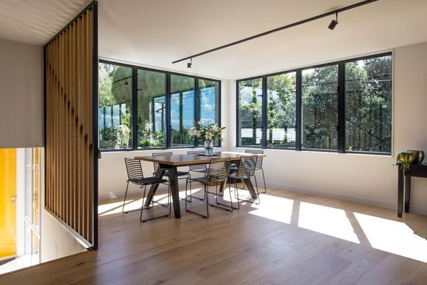 Bright contemporary dining area with a timber dining table and black wire-frame chairs, set against expansive black-framed windows. A vertical timber slat partition partially screens the adjoining stair or lower-level opening, while track lighting and pale timber flooring create an airy, minimalist interior.
