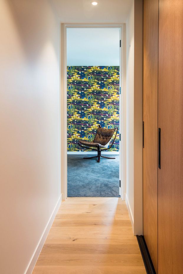A narrow, softly lit residential hallway with pale timber flooring, white walls, and tall warm-wood built-in cabinetry along the right side. The hallway opens into a carpeted lounge or living area featuring a brown leather lounge chair and a bold multicoloured patterned feature wall.