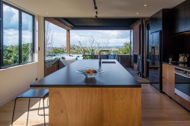 A contemporary kitchen centred on a large timber-fronted island with a dark benchtop and integrated cooktop. Full-height windows and wide open glazing connect the kitchen to a covered outdoor entertaining area and leafy coastal landscape. Dark built-in cabinetry, timber flooring, track lighting and bar stools complete the open-plan space.