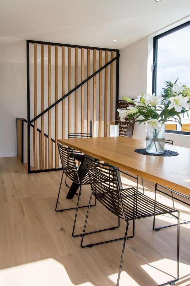 Bright contemporary dining area with a long natural timber dining table and black wire-frame chairs. A distinctive black-framed timber slat stair balustrade forms a strong architectural feature, with pale timber flooring, white walls and ceiling, and large black-framed window glazing bringing in abundant daylight.