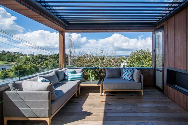 A bright, enclosed outdoor lounge or deck with timber flooring, timber-clad walls, extensive glazing, and a slatted overhead pergola. It contains a grey sectional sofa, armchair, coffee table, cushions, potted greenery, and a built-in dark fireplace or television recess. The space overlooks a leafy suburban landscape and distant water or hills.