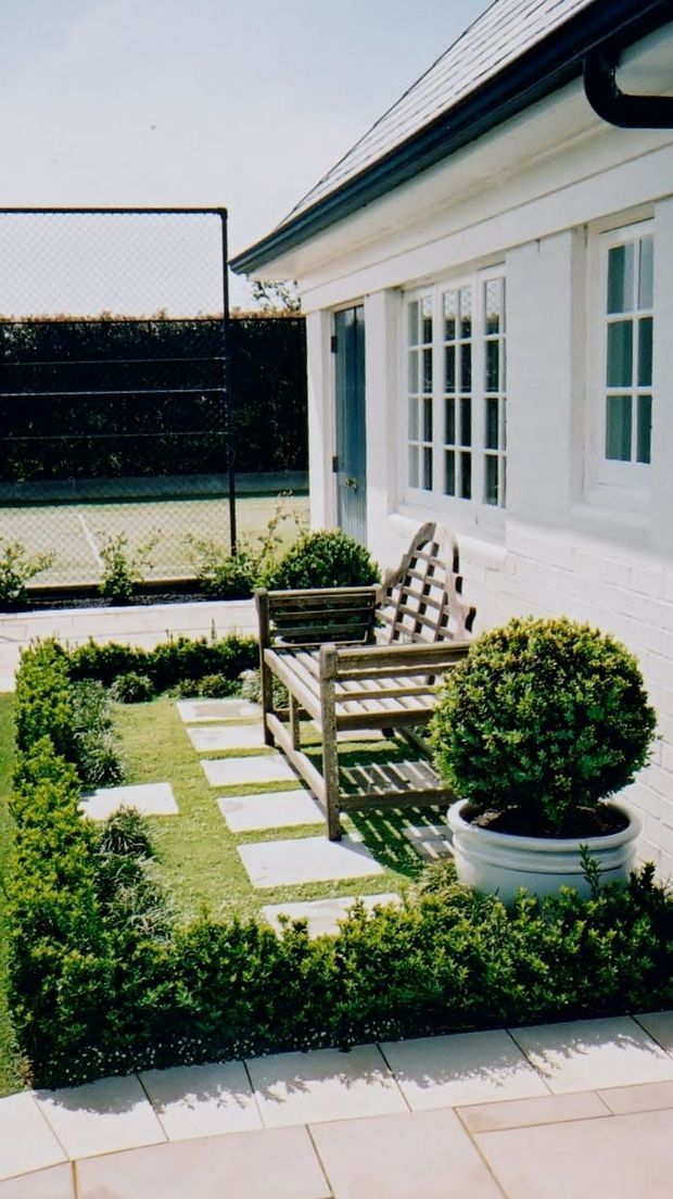 A bright, neatly landscaped courtyard beside a white-painted brick building. The building has multi-pane white-framed windows, a glazed door, a pitched roof with dark gutters, and a small timber bench on a paved garden path. Clipped shrubs, a large potted topiary, lawn, pale stepping stones, and a chain-link sports court are visible.
