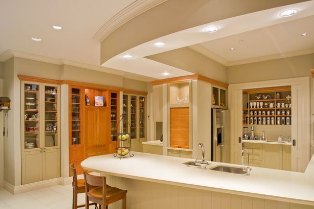 A spacious, warm-toned traditional kitchen with extensive cream built-in cabinetry, glass-front display cupboards, a large white island with seating, integrated appliances, and a raised serving or viewing opening. The room features decorative ceiling curves, recessed lighting, timber cabinetry accents, and a well-organised beverage or display area.