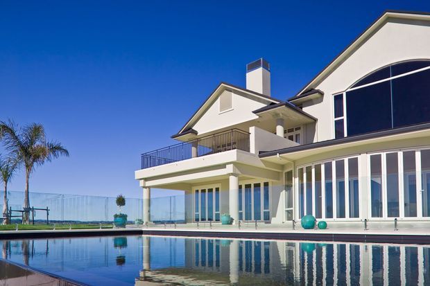 Exterior view of a large white multi-storey residence beside a swimming pool. The home has a prominent gabled roof, projecting balconies, columns, extensive floor-to-ceiling glazing, and a curved glazed facade section. Palm trees and a fenced outdoor area are visible around the property, with strong reflections in the pool.