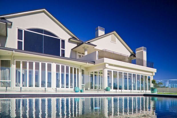 Exterior view of a large multi-level residential house beside a swimming pool. The home has pale rendered walls, pitched gable roofs, broad white-framed glazing, a curved upper-level window, balconies with metal balustrades, and an enclosed sunroom-like ground-floor terrace. The pool dominates the foreground and reflects the building.