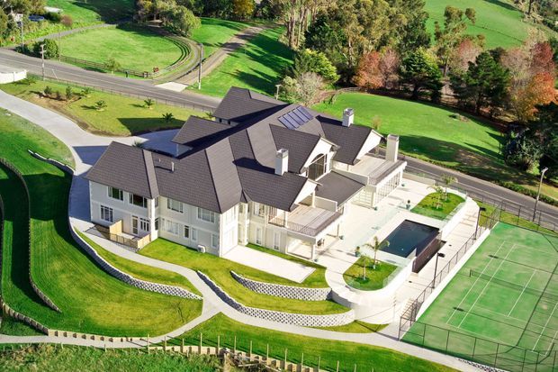 Aerial view of a large, multi-wing residential estate set within expansive landscaped grounds. The light-coloured house has a complex dark pitched roof, multiple gables, dormer-like projections, balconies and terraces. A swimming pool, paved outdoor entertaining area and full-size tennis court are visible beside the home, with curving paths and manicured lawns surrounding the property.