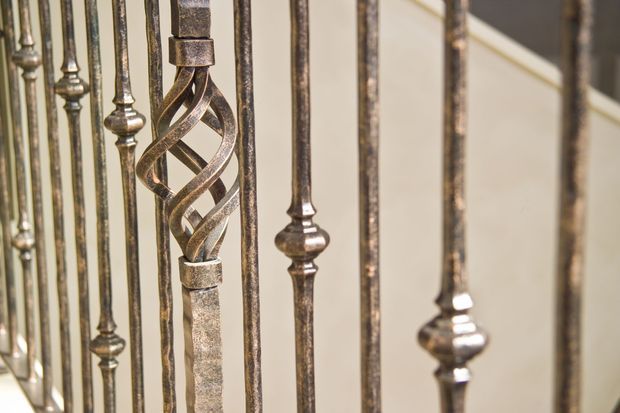 Close-up detail of ornate wrought-iron stair balustrading, featuring slender vertical metal spindles with decorative turned sections and a prominent twisted central motif. A pale stair or wall surface is visible behind the railing.