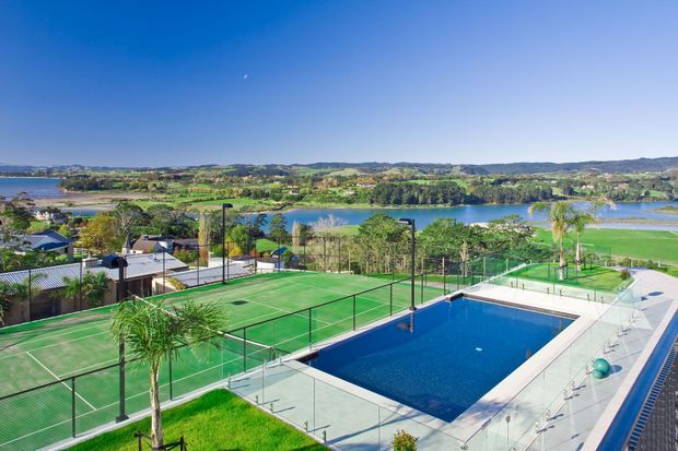 Elevated exterior view of a landscaped recreational property featuring a blue swimming pool, multiple fenced green tennis courts, paved surrounds, and palm trees. A broad lake and rolling green landscape are visible beyond the site under a clear blue sky.