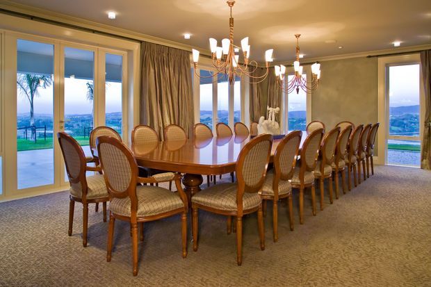 Formal dining room with a long polished wooden dining table surrounded by numerous cane-backed upholstered dining chairs. The room features patterned carpet, full-height glazed doors and windows with heavy drapery, warm beige walls, and multiple ornate chandelier-style pendant lights. A scenic landscape is visible outside.