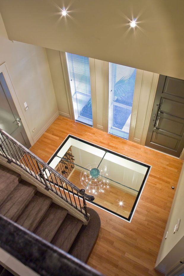 A bright multi-level residential stairway and landing viewed from above. The space features warm timber flooring, carpeted stairs with dark ornate metal balustrades, tall glazed doors or windows with horizontal blinds, recessed ceiling lights, and a glass floor opening over a decorative chandelier below.