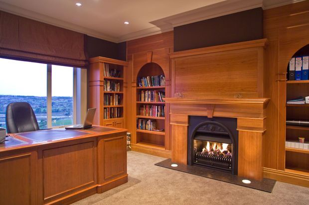 A richly wood-panelled home office with a substantial built-in timber desk positioned beside a large window, extensive built-in bookshelves, a concealed television cabinet above a traditional fireplace, and a gas fire. Warm recessed lighting, dark window blinds, patterned carpet, and an expansive landscape view create a refined, traditional study atmosphere.