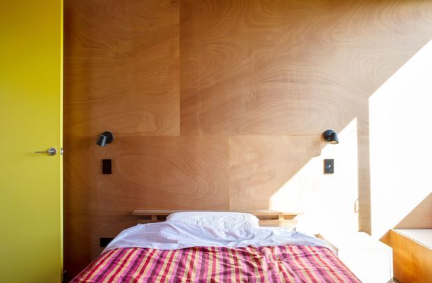 A compact bedroom with a bed positioned against a full-height warm timber feature wall. The wall incorporates small black reading lights and electrical switches, with narrow timber bedside ledges. A vivid lime-green door contrasts with the plywood-toned wall, while strong sunlight enters from the right and falls across the room. The bed has white bedding and a brightly striped pink, red, orange, and green cover.