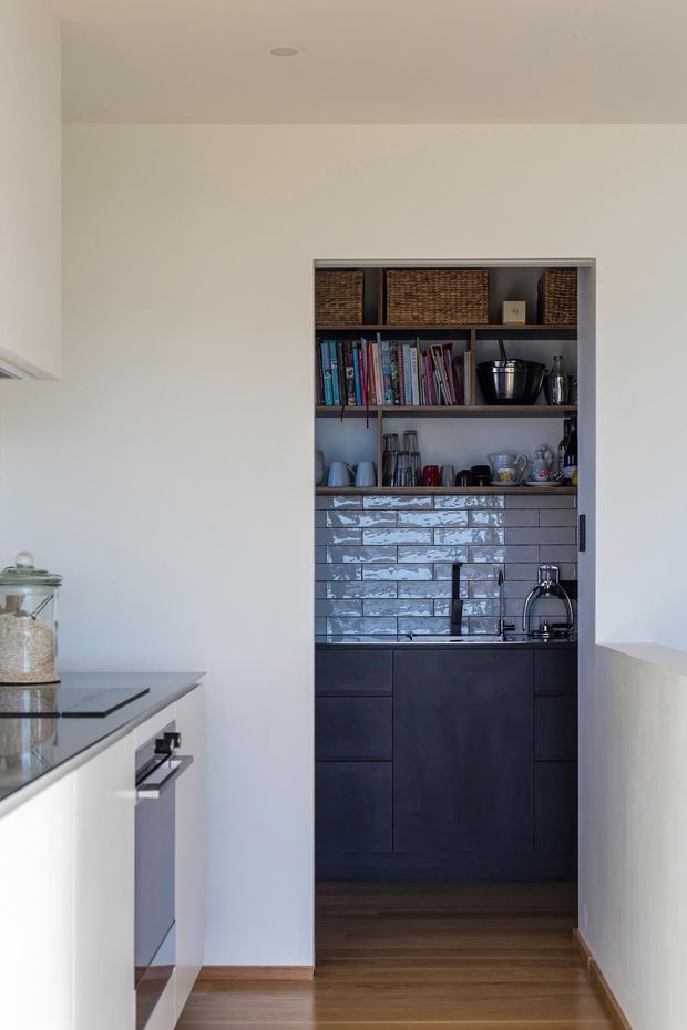 A narrow, refined kitchen view focused on a dark built-in butler's pantry or kitchenette. The recessed joinery has open shelving with books, woven baskets, glassware and cookware above dark cabinetry, a black sink and tap, and a glossy grey subway-tile splashback. In the foreground, a pale kitchen island with a dark stone benchtop is partially visible. Warm timber flooring and white walls create a clean, contemporary interior.
