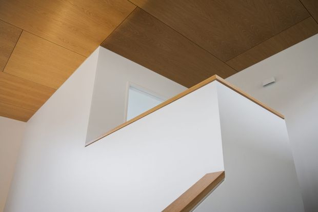 Upward-looking interior view of a contemporary stairway or landing, featuring crisp white walls and stair balustrades edged with light timber trim. A warm timber-clad ceiling creates a strong geometric canopy above, with a partially visible doorway or opening behind the stair structure.