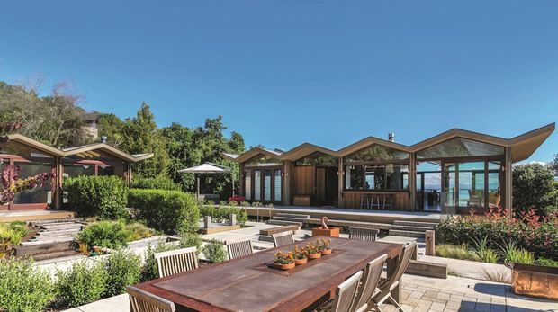 A bright exterior view of a low-rise contemporary residence arranged around landscaped terraces and a large outdoor dining area. The building has angular, multi-gabled roof forms, extensive floor-to-ceiling glazing, timber-toned framing, and stepped paved garden spaces. A glimpse of blue water is visible beyond the glazing, while planted hedges and garden beds frame the courtyard-like setting.
