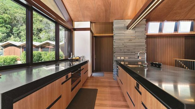 A warm mid-century-inspired kitchen with extensive timber cabinetry, dark benchtops, and a central island. Floor-to-ceiling glazing along one side overlooks lush greenery and small outbuildings. A stacked natural-stone chimney or feature wall anchors the rear of the room, while linear pendant lighting and vertical timber wall panelling reinforce the strong horizontal and vertical geometry.