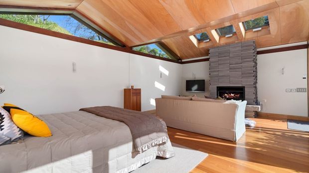 A bright, open-plan bedroom with a large bed in the foreground and a sitting area beyond. The room features warm timber flooring and a dramatic timber-lined sloped ceiling with clerestory glazing and multiple skylights. White walls, a freestanding timber cabinet, a television, and a stacked grey masonry fireplace with an inset fire create a modern natural interior. Strong sunlight enters through the roof glazing, highlighting the spacious, airy character.