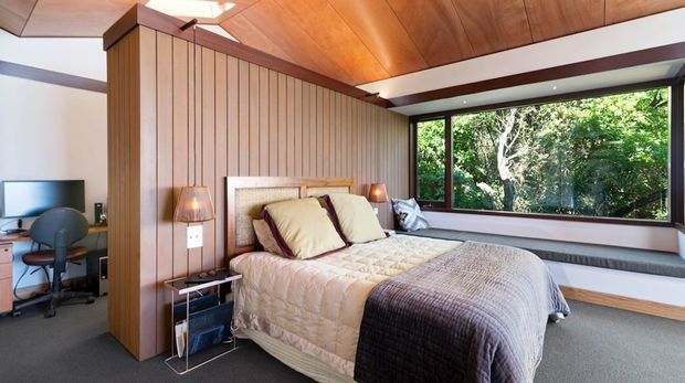 A warm, mid-century-inspired bedroom with a bed positioned against a full-height vertical timber-panel feature wall. The room has a richly timber-lined sloping ceiling with dark exposed beams, integrated bedside lighting, a compact desk and office nook, and a broad window seat beneath expansive dark-framed glazing overlooking greenery. Neutral bedding and dark carpet create a calm, cosy interior.
