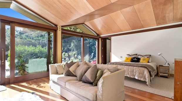 Bright bedroom or studio-like sleeping space with a large upholstered sofa in the foreground and a bed behind it. The room features extensive timber-framed glazing, a warm timber-lined sloping ceiling with exposed beams, timber flooring, and views onto lush vegetation. Neutral upholstered furnishings and bedding create a relaxed, light-filled atmosphere.