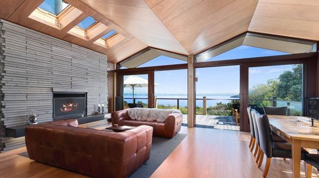 A spacious open-plan living and dining room with expansive glazing overlooking a coastal or waterfront setting. The room features a dramatic angled timber-lined ceiling with multiple skylights, a large horizontal stacked-stone fireplace wall, warm timber flooring, leather sofas, and a substantial timber dining table. Structural timber posts and exposed roof geometry create a strong contemporary architectural character.