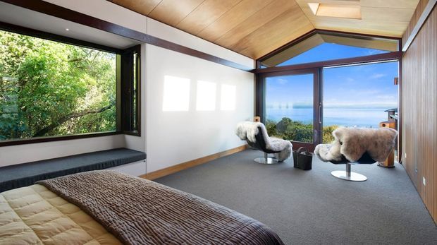 A spacious bedroom with a low-profile bed in the foreground, charcoal carpet, and two sculptural lounge chairs positioned beside expansive glazing. The room features warm timber ceiling and wall lining, dark timber window and door frames, and a dramatic angled ceiling form. Large windows and glazed doors open the room visually to lush vegetation on one side and a bright ocean or coastal outlook beyond.
