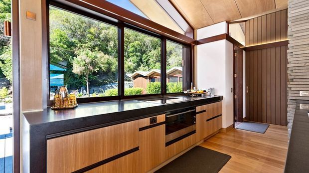 A compact, contemporary kitchen with extensive floor-to-ceiling glazing overlooking a lush landscaped setting. The room features warm timber cabinetry and ceiling panels, a long dark stone benchtop, integrated black appliances, dark-framed windows, and a vertically slatted timber wall near the entry. Natural light and greenery create a warm indoor-outdoor connection.