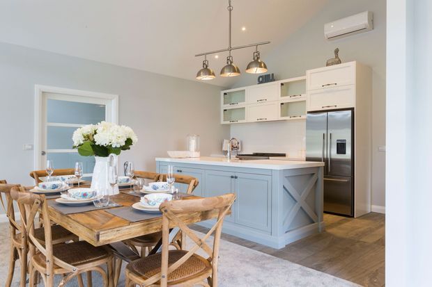 Bright open-plan kitchen and dining area with a pale blue island and cabinetry, white benchtop, stainless-steel refrigerator, open shelving, and industrial-style pendant lights. A rustic timber dining table with woven chairs is set for a meal, creating a warm coastal farmhouse atmosphere. Light grey walls, timber-look flooring, and a large rug complete the space.