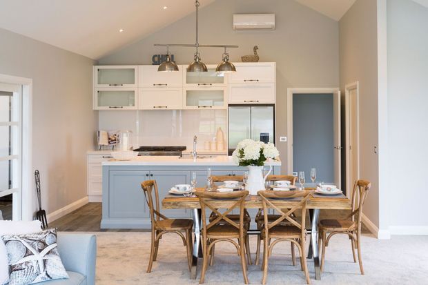 Bright open-plan kitchen and dining area with a pale blue island, white built-in cabinetry, light stone benchtops, pendant lights, and a timber dining table with woven-style chairs. The room has a vaulted ceiling, pale grey walls, carpeted flooring, and a relaxed, refined coastal-country feel.