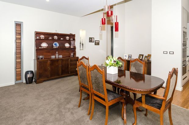 A bright formal dining room with a polished dark timber dining table and upholstered carved timber chairs. A traditional timber display hutch occupies the left wall, while a contemporary cluster of red pendant lights hangs above the table. The room has white walls, grey carpet, framed artwork, and an open connection to a timber-floored area beyond.