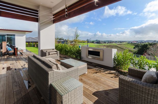 Covered outdoor entertaining terrace with timber decking, woven outdoor lounge furniture, a glass coffee table, built-in fireplace, and adjustable timber screening. The terrace overlooks a landscaped garden and open suburban/rural surroundings under a bright sky.