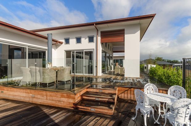 Exterior view of a contemporary multi-level home with a broad timber deck and stairs, glazed balustrades, white rendered walls, projecting rooflines, and covered outdoor living areas. The deck includes outdoor dining furniture and lounge seating, with suburban surroundings visible beyond.