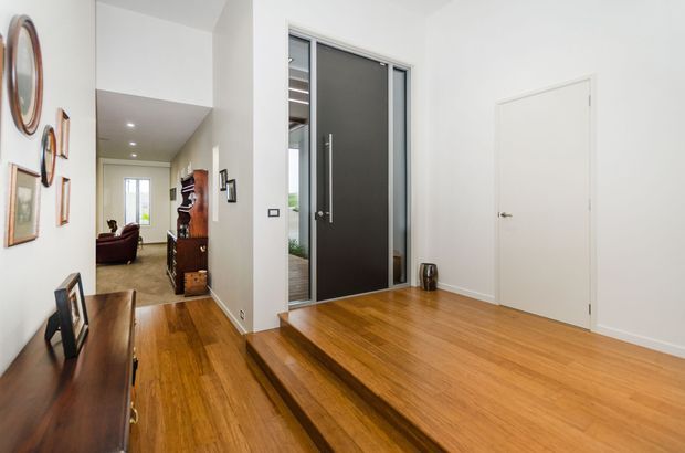 Bright contemporary entry foyer with warm timber flooring, a raised landing and steps, a dark-framed glazed entry door, white walls, and a hallway leading toward a living area. Framed artwork and a timber console add understated decoration.