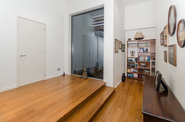 A bright interior hallway or landing with polished timber flooring and two raised steps leading to a lower-level passage. White walls and doors create a clean backdrop, while a full-height glazed opening reveals another room or stairwell. A built-in timber bookshelf, dark console desk, framed artwork, mirrors, and decorative objects add warmth and character.