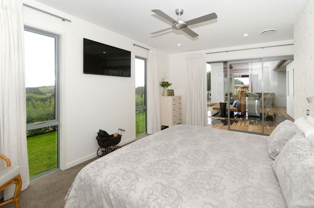 Bright contemporary bedroom with a large bed, white patterned bedding, floor-to-ceiling windows, sheer curtains, wall-mounted television, ceiling fan, and a sliding glass opening to a dining area beyond. The room has views toward a green rural landscape and a neutral white, grey, and light timber palette.
