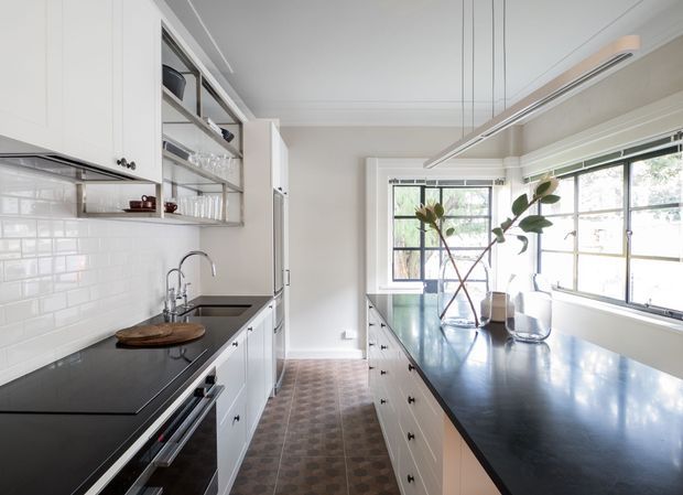 Bright, classic galley-style kitchen with white shaker cabinetry, dark stone or composite benchtops, a white subway-tile splashback, and a patterned tiled floor. The kitchen includes a long central island with extensive drawers, integrated cooking appliances, open shelving with glassware, and a large bank of windows providing abundant natural light.