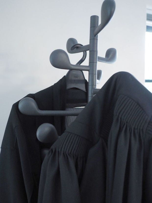 Close-up photograph of a dark grey freestanding coat rack with multiple rounded hooks, holding several black coats and garments. The rack is positioned against a bright white interior wall, with a small portion of a window visible in the background.