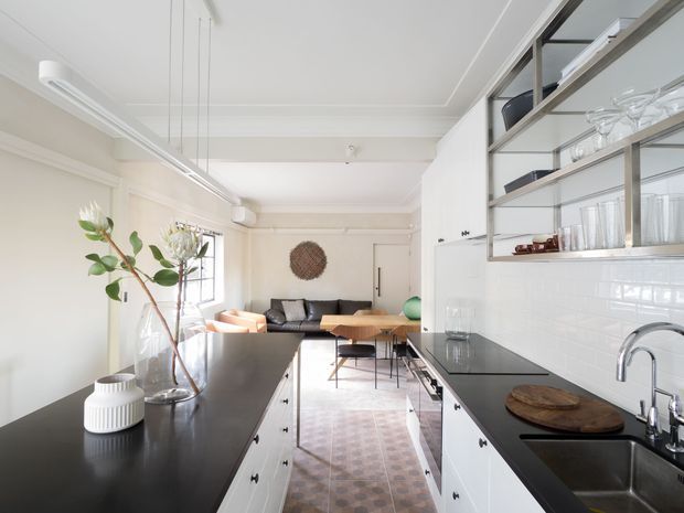 Bright, narrow open-plan kitchen with white shaker-style cabinetry, black stone benchtops, glossy white subway-tile splashback, and open metal-framed shelving. The kitchen opens directly toward a compact living area with a dark sofa, timber coffee table, and patterned rug. Large windows and a light neutral palette create an airy, contemporary feel.