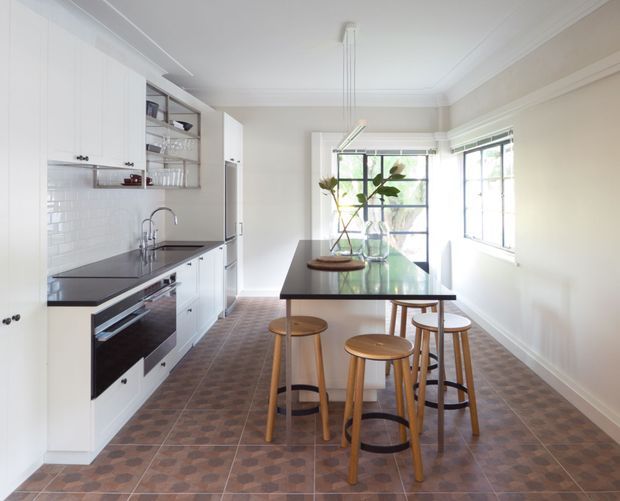 Bright, narrow kitchen with a long central island, integrated seating, white shaker-style cabinetry, dark benchtops, white subway-tile splashback, and large gridded windows. The room has decorative cornices and a patterned brown tiled floor, combining contemporary functionality with traditional detailing.