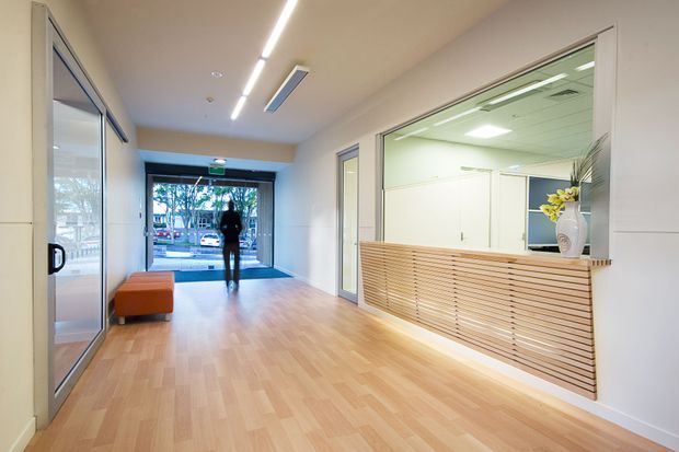 Bright contemporary commercial entry lobby or reception area with pale timber flooring, white walls, linear ceiling lighting, glazed office or reception spaces, and a built-in slatted timber reception counter with concealed underlighting. A glass entrance opens to the street, and a small orange bench provides seating.