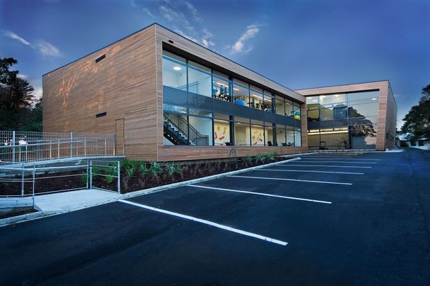 Exterior view of a modern two-storey commercial building with extensive floor-to-ceiling glazing, warm horizontal timber cladding, exposed internal staircases, and a raised accessible walkway. The building fronts a marked asphalt parking area and is photographed at dusk under a blue sky.