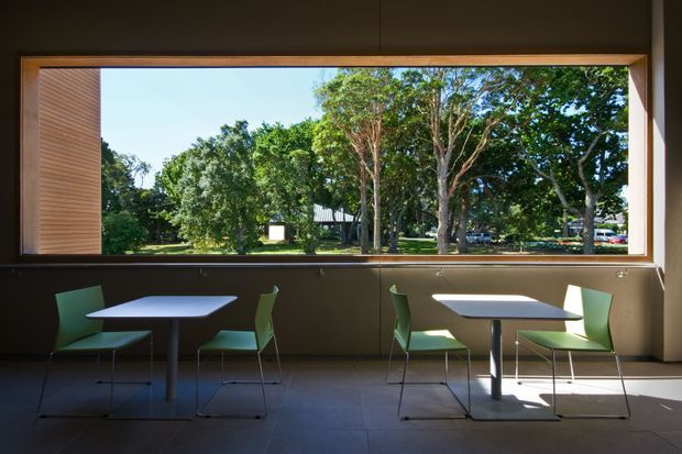 A modern interior dining or casual seating area with two small square tables and green chairs arranged beside a large horizontal picture window. Dark brown wall panels and a tiled floor frame views of leafy grounds outside, with strong daylight creating a calm, understated atmosphere.