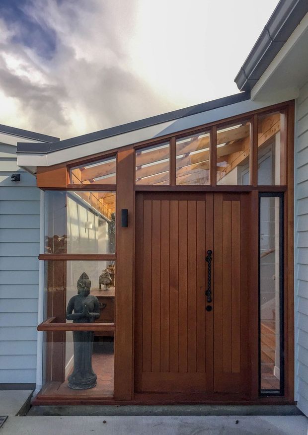 Exterior view of a residential entryway featuring a tall timber front door with vertical paneling and dark hardware, surrounded by extensive timber-framed glazing. The entry is set within pale blue-grey horizontal weatherboard cladding, with exposed timber roof structure visible through the glazing and a low-pitched roofline.