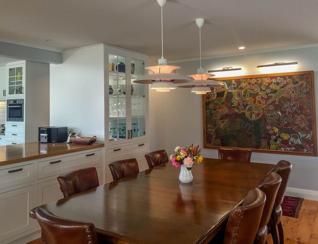 A warm, refined dining room with a large dark timber dining table surrounded by brown leather chairs. Built-in light painted cabinetry and glass-front display cupboards line the left side, while a large colourful framed artwork forms the main feature wall. Multiple pendant lights hang above the table, complemented by discreet wall lighting. Timber flooring and neutral walls create a traditional, inviting atmosphere.