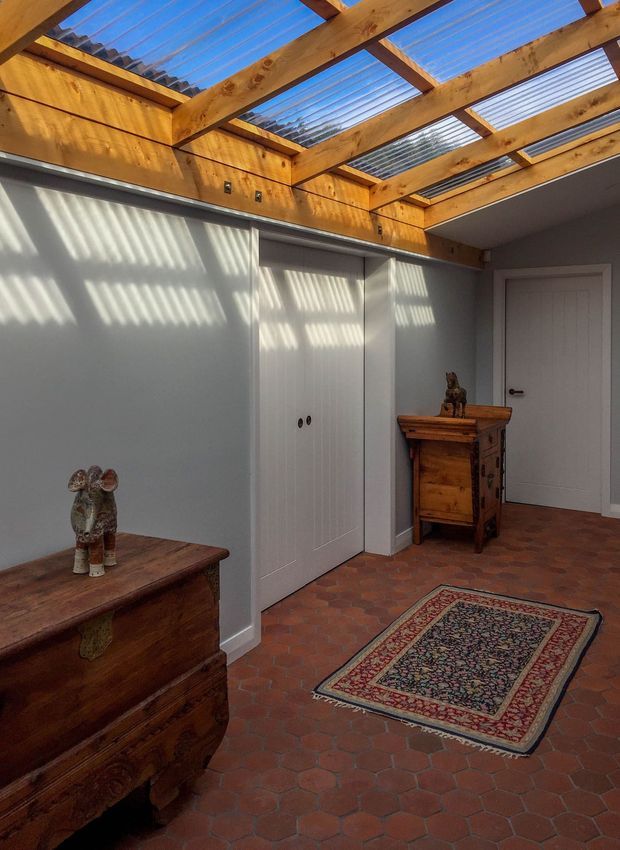 A compact interior hallway or entry area with white built-in storage cupboards, a white hinged door, warm timber exposed roof beams and translucent corrugated roof panels creating striped sunlight, and a reddish terracotta hexagonal tile floor. Antique wooden chests and a patterned runner add traditional character.