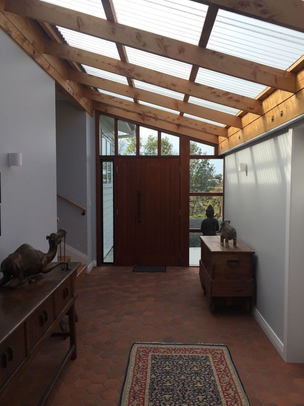 A narrow, light-filled entry hall with a dramatic timber-and-translucent-panel overhead roof structure. A dark timber front door with glazed sidelights and transom opens onto an exterior view. The hall has pale painted walls, warm terracotta hexagonal floor tiles, timber furniture, a patterned runner, wall sconces, and decorative objects. Exposed timber beams and clerestory-like glazing create a warm, rustic architectural character.