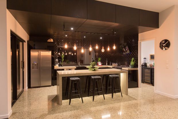 A spacious contemporary kitchen with a large stone island and three dark bar stools, extensive dark overhead cabinetry, stainless-steel refrigerator, black brick splashback, and a row of warm pendant lights. The polished speckled floor and dark ceiling create a dramatic, refined atmosphere, with an adjacent dining or living area visible beyond the kitchen.